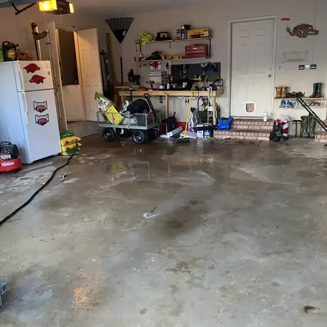 Water spread across a garage floor with hoses and cleanup equipment near stored household belongings